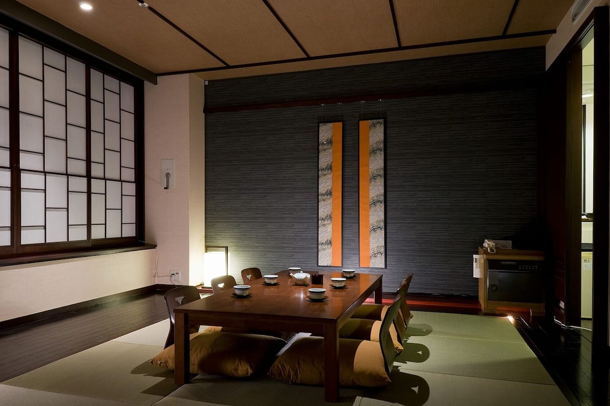 Une pièce de style japonais traditionnel, avec un sol recouvert de tatamis, une petite table basse avec un service à thé, des portes coulissantes et un mur sombre à la texture marquée.