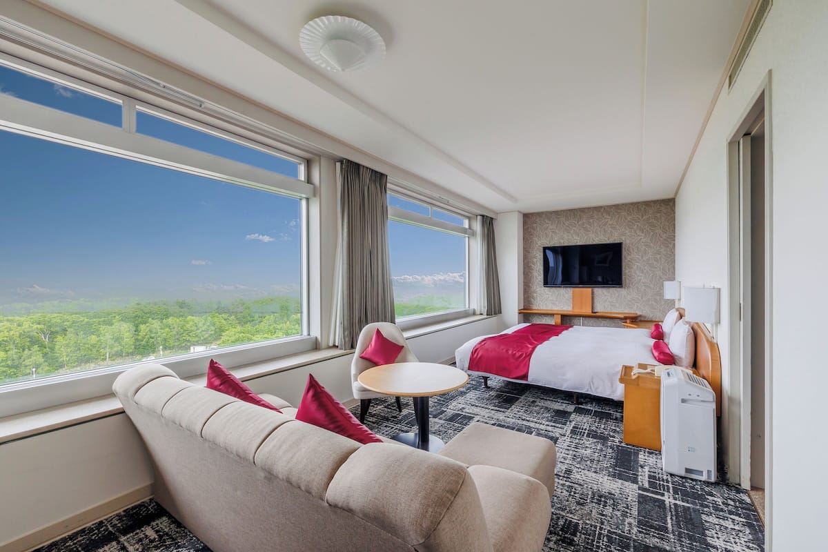 Une chambre d’hôtel avec un lit, un canapé, une petite table, une télévision et une fenêtre donnant sur un paysage verdoyant.