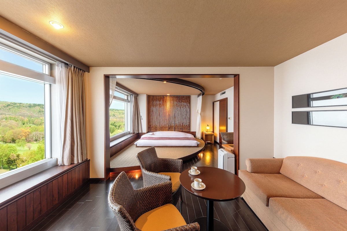 Une chambre d’hôtel avec un lit, un canapé, une table à manger avec des tasses, et une vue sur la verdure à l’extérieur.