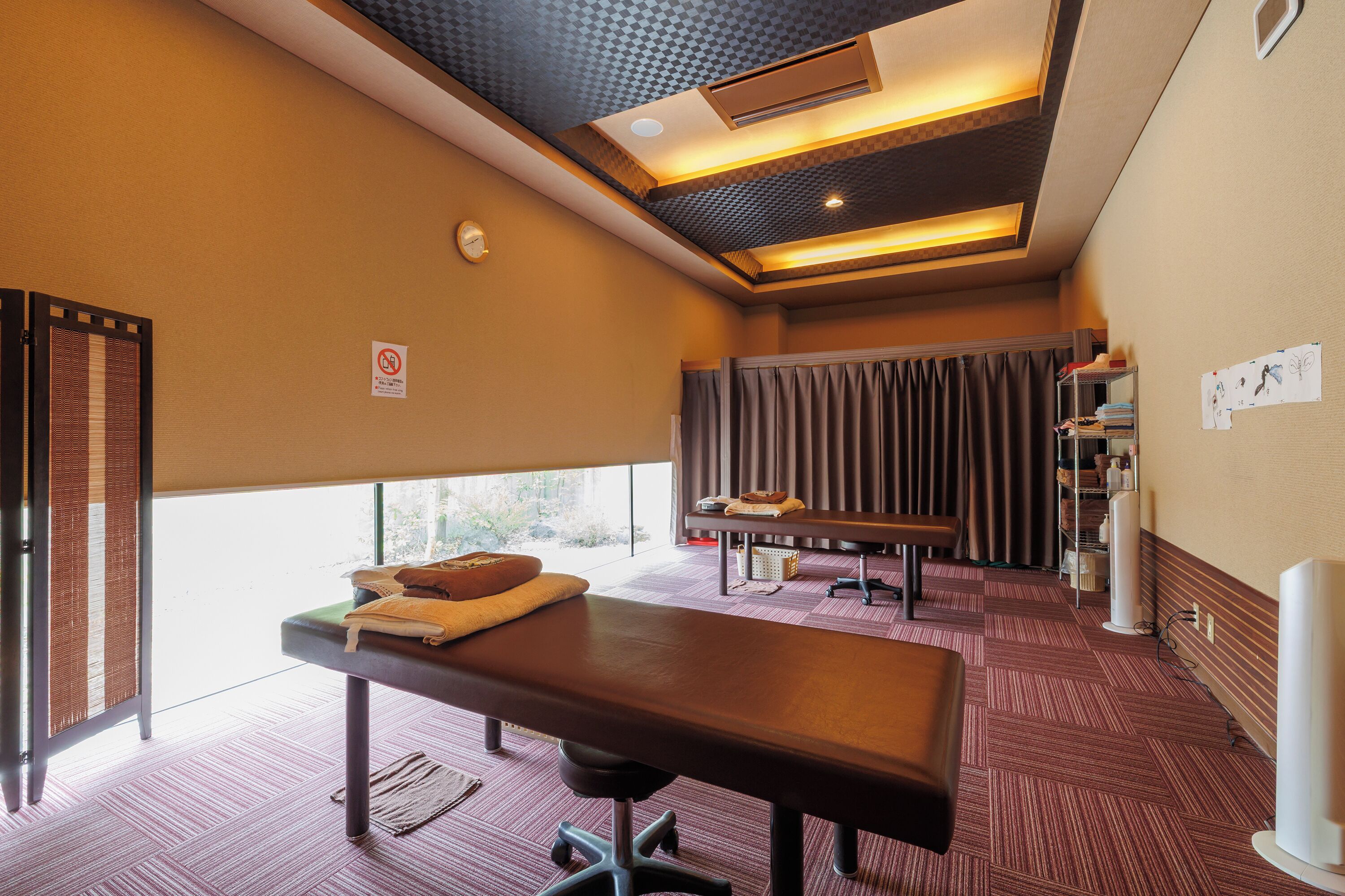 A room with a massage table, a bench, a window with curtains, and a shelf with books and bottles.