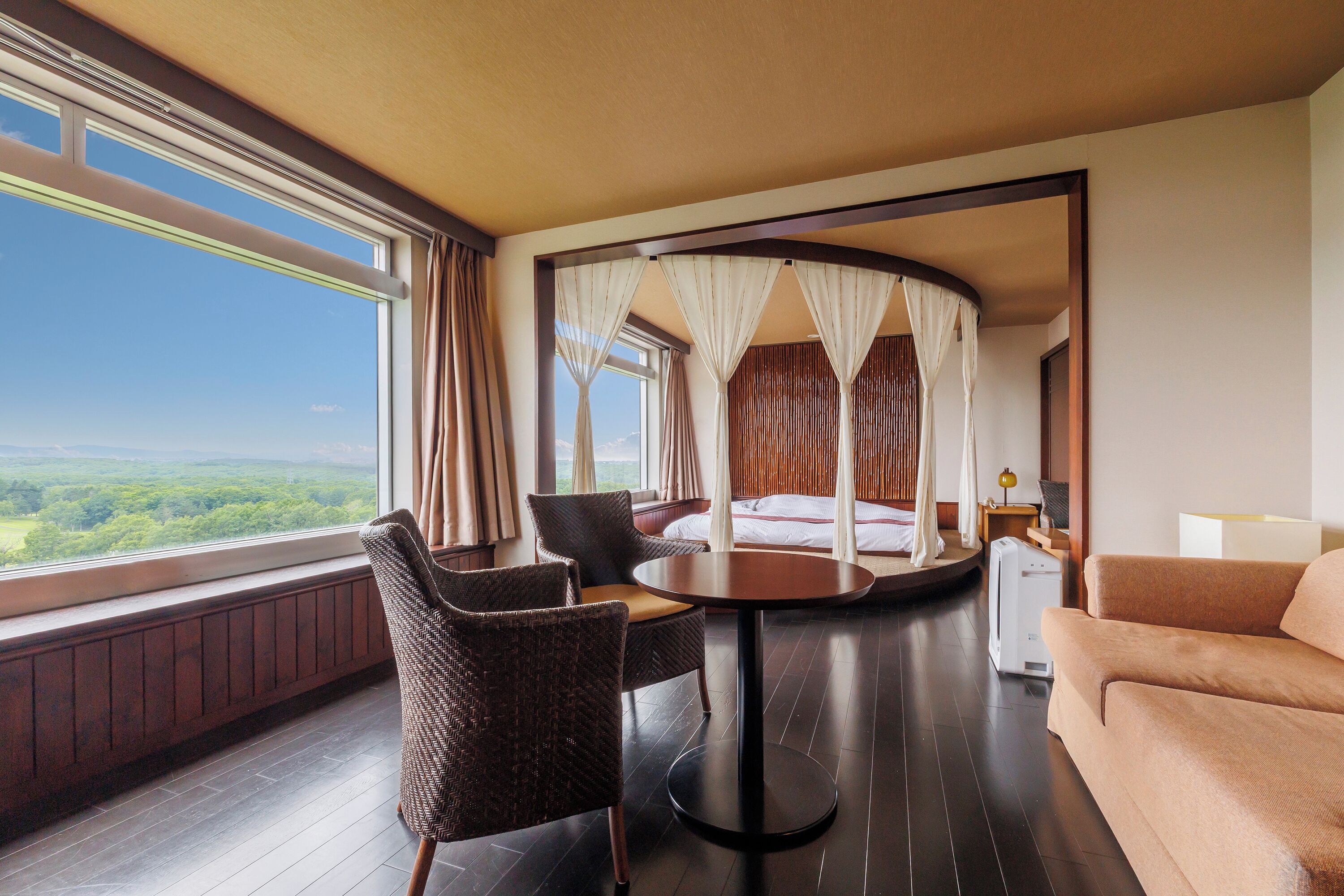 A hotel room with a round table, two chairs, a sofa, a bed with a canopy, and a view of the outdoors.