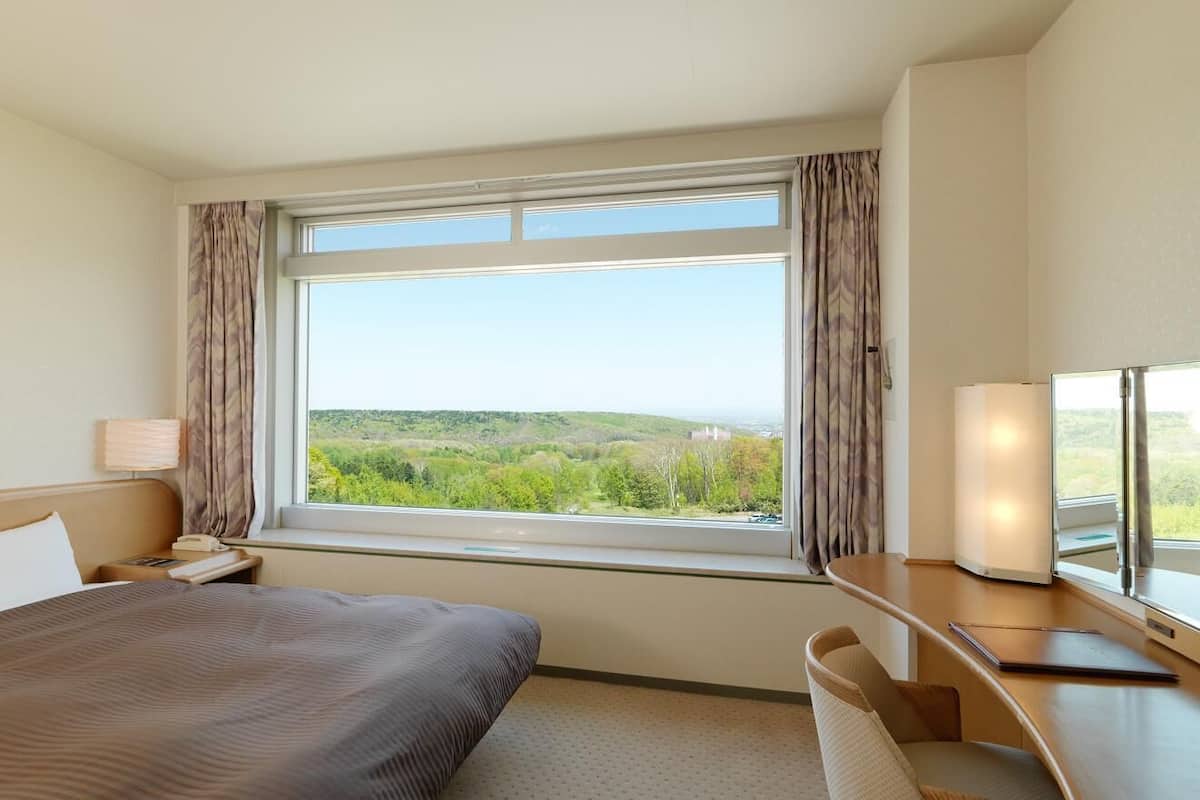 Une chambre d’hôtel équipée d’un lit, d’un bureau, d’une chaise, d’une table de chevet, d’une lampe et d’une fenêtre donnant sur un paysage verdoyant.