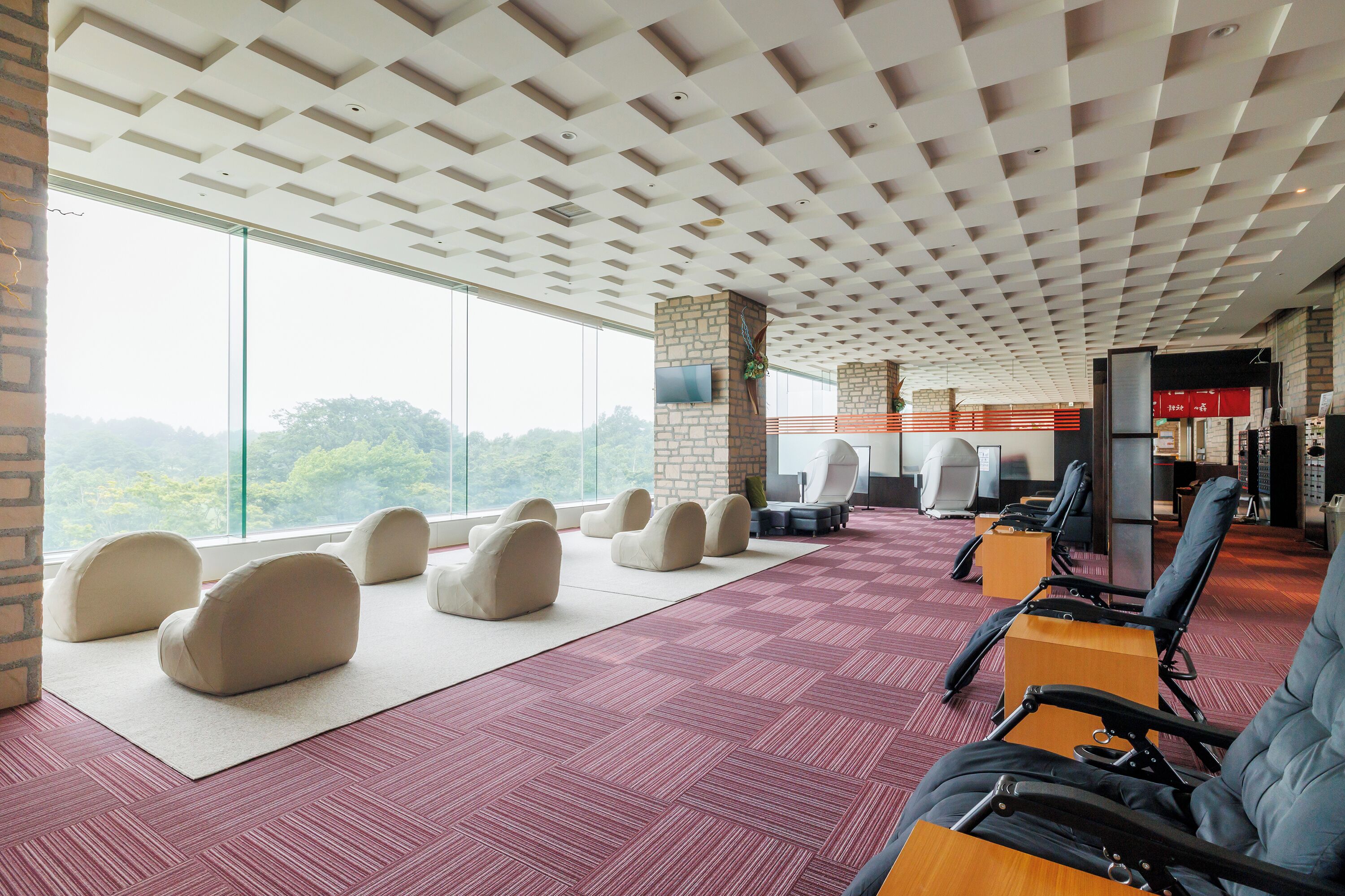 A spacious room with a high ceiling, large windows, and a view of greenery. There are several seating areas with chairs and a massage table.