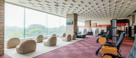 A spacious room with a high ceiling, large windows, and a view of greenery. There are several seating areas with chairs and a massage table.