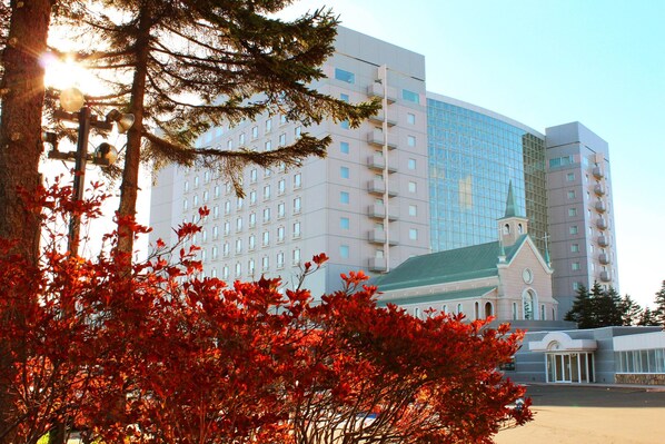 Un hotel de varios pisos con fachada de vidrio moderna, un edificio con techo verde que recuerda a una iglesia, y un estacionamiento en primer plano.