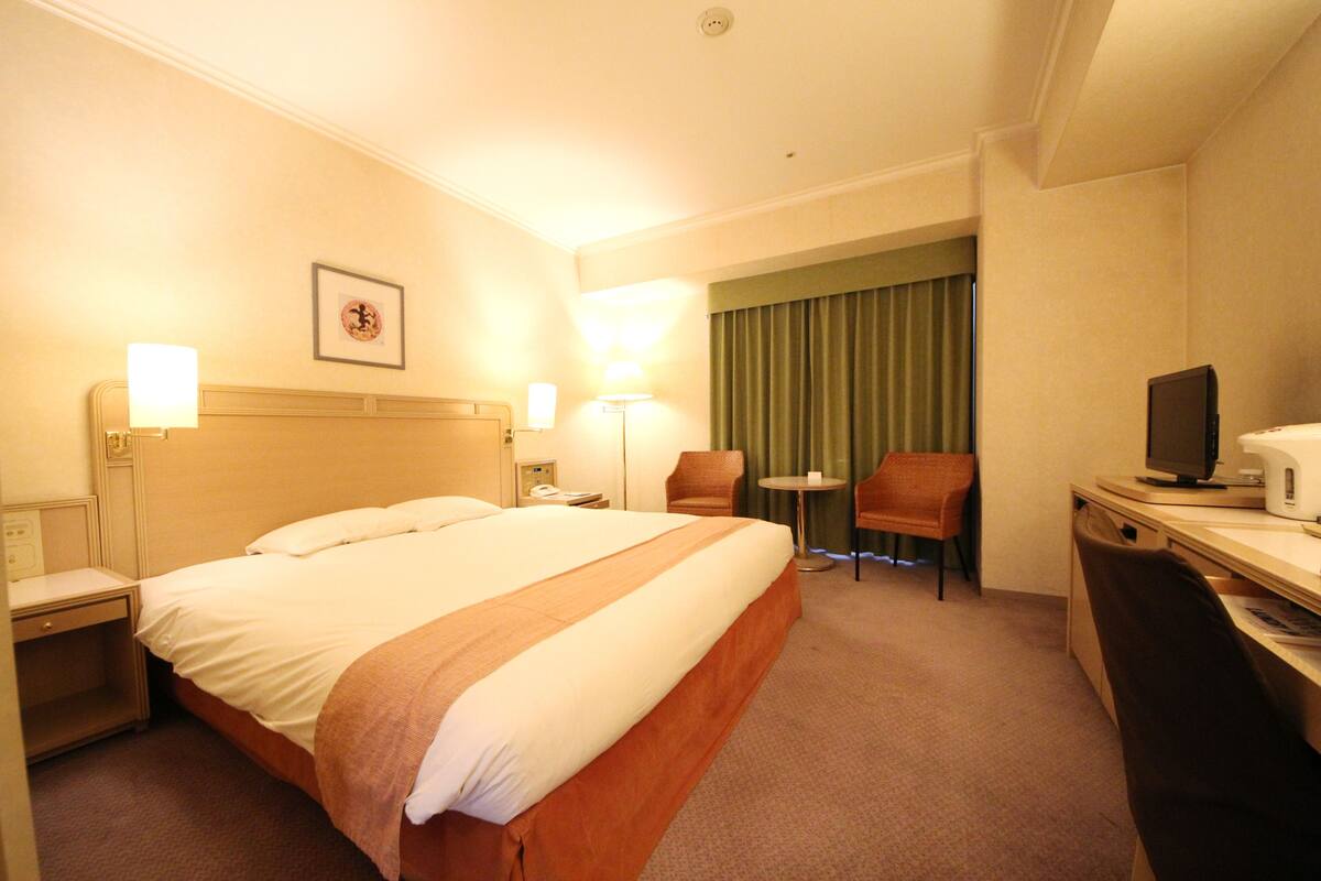 Habitación de hotel con cama, mesitas de noche, escritorio, silla, lámpara y televisor.