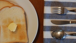 Une assiette avec des tartines beurrées, un verre contenant un liquide sombre et une serviette avec des couverts, le tout posé sur une table en bois.