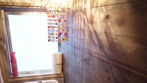 Une petite salle de bain avec un mur en bois, des toilettes blanches, un petit lavabo et une fenêtre agrémentée d’une décoration colorée.