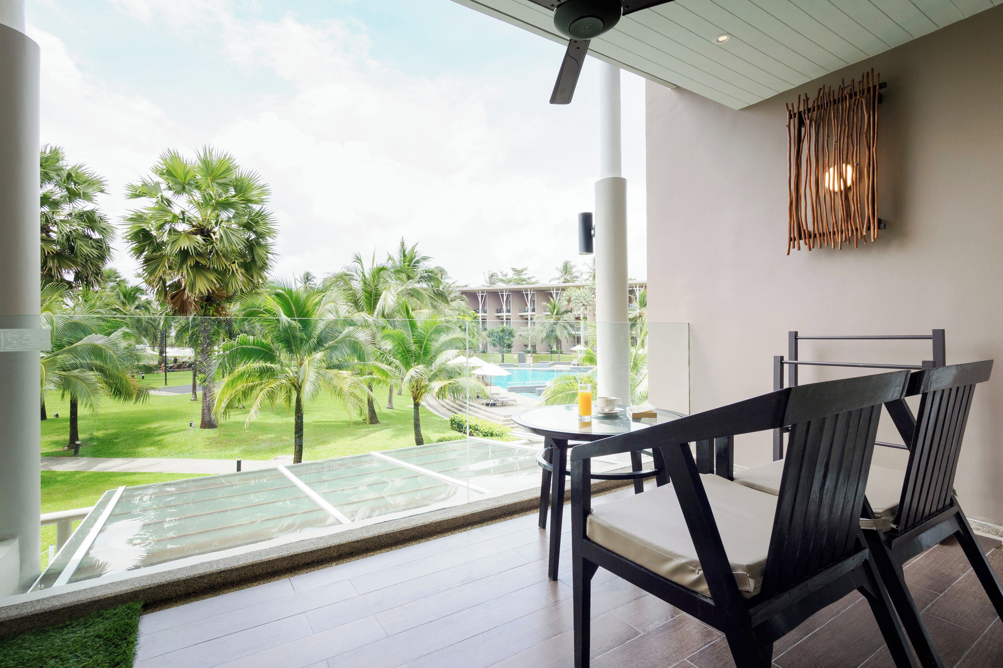 Un balcón con piso de vidrio, una mesa y sillas, con vista a una piscina y palmeras.