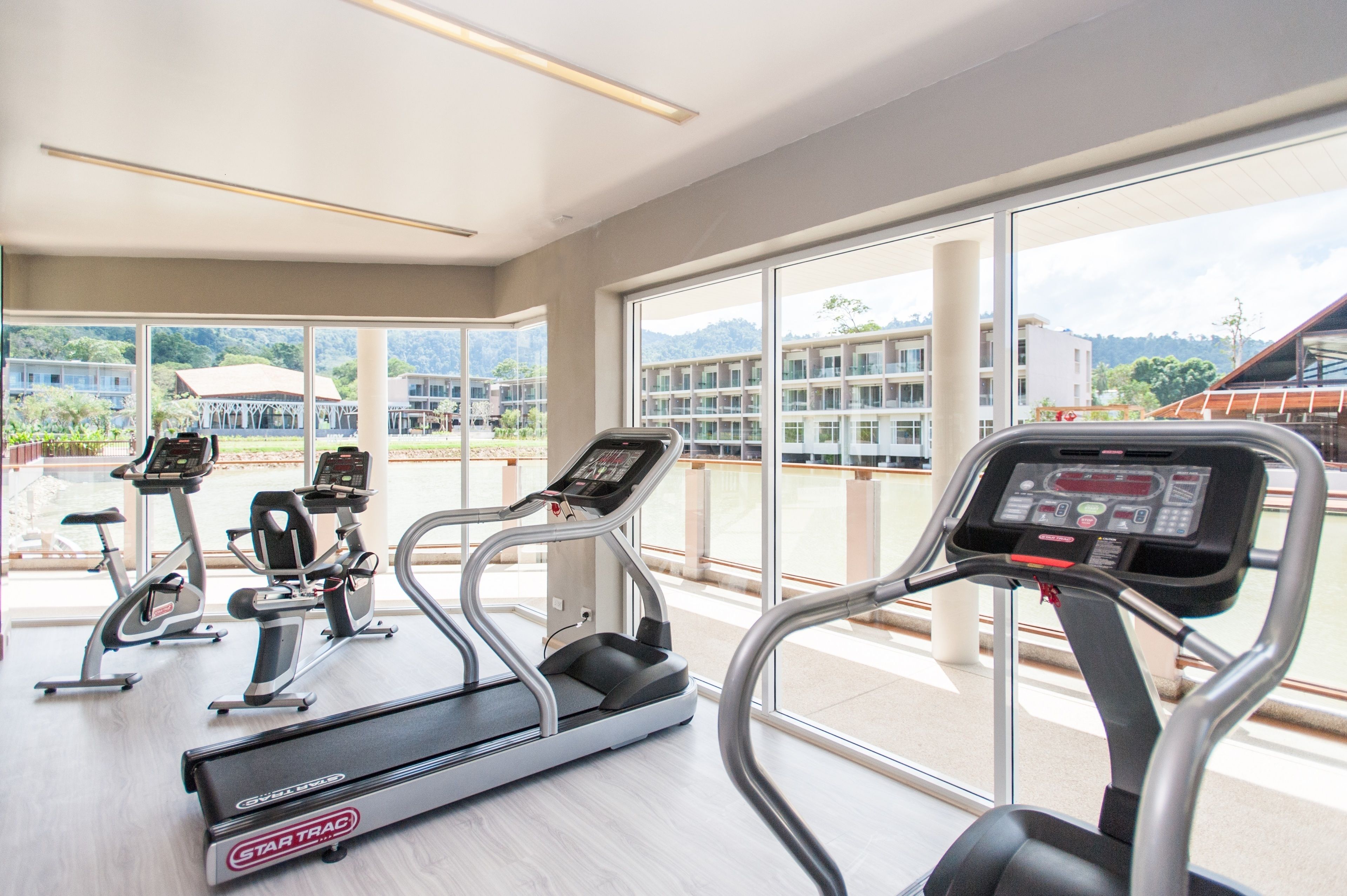 A gym with treadmills, ellipticals, and stationary bikes, large windows with a view of buildings and greenery.