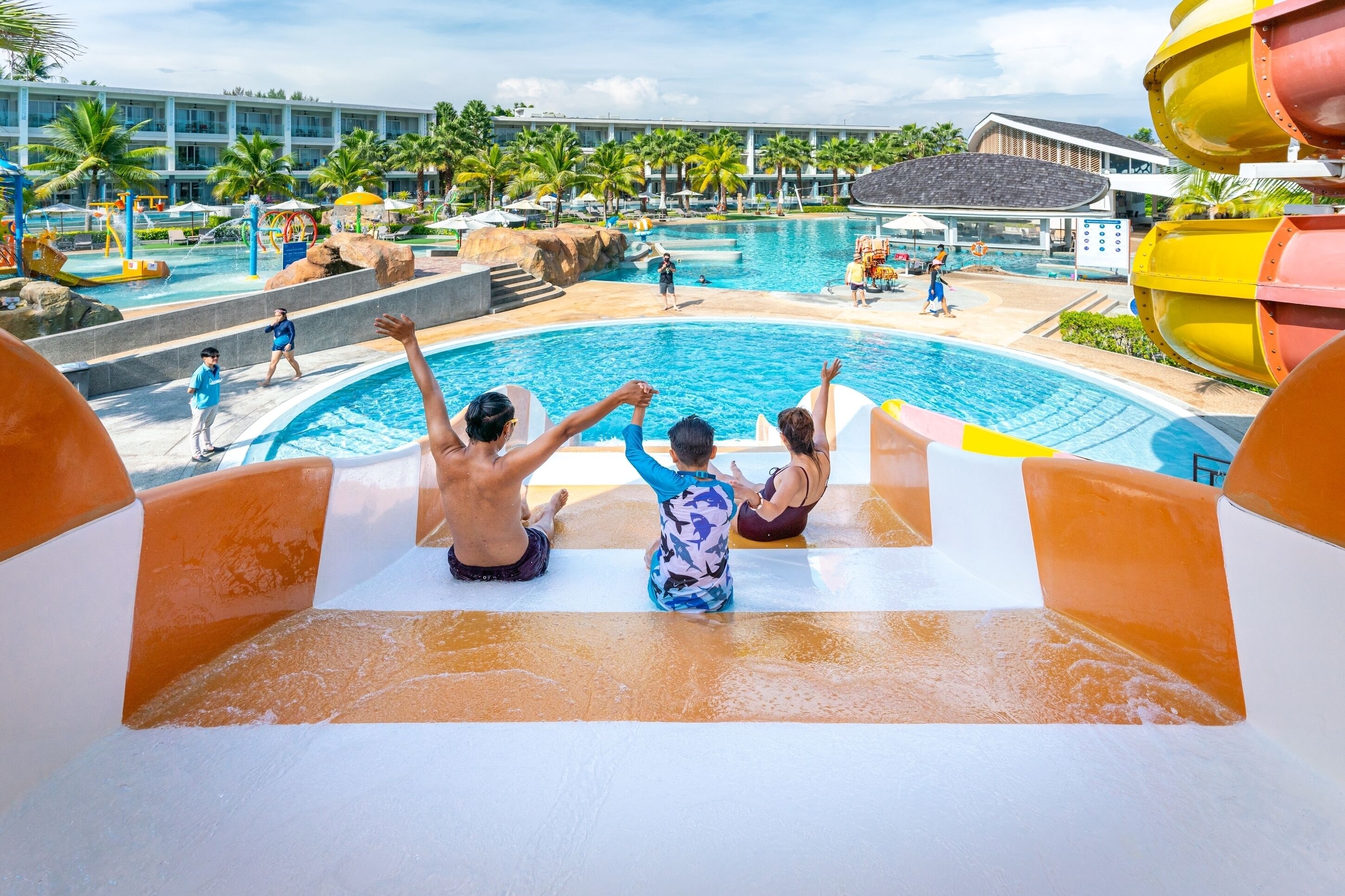 Un toboggan aquatique qui débouche sur un espace piscine agrémenté de palmiers et avec un bâtiment en arrière-plan.