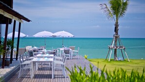 A wooden deck with white chairs and tables, overlooking a clear blue sea, with two striped umbrellas and a palm tree.