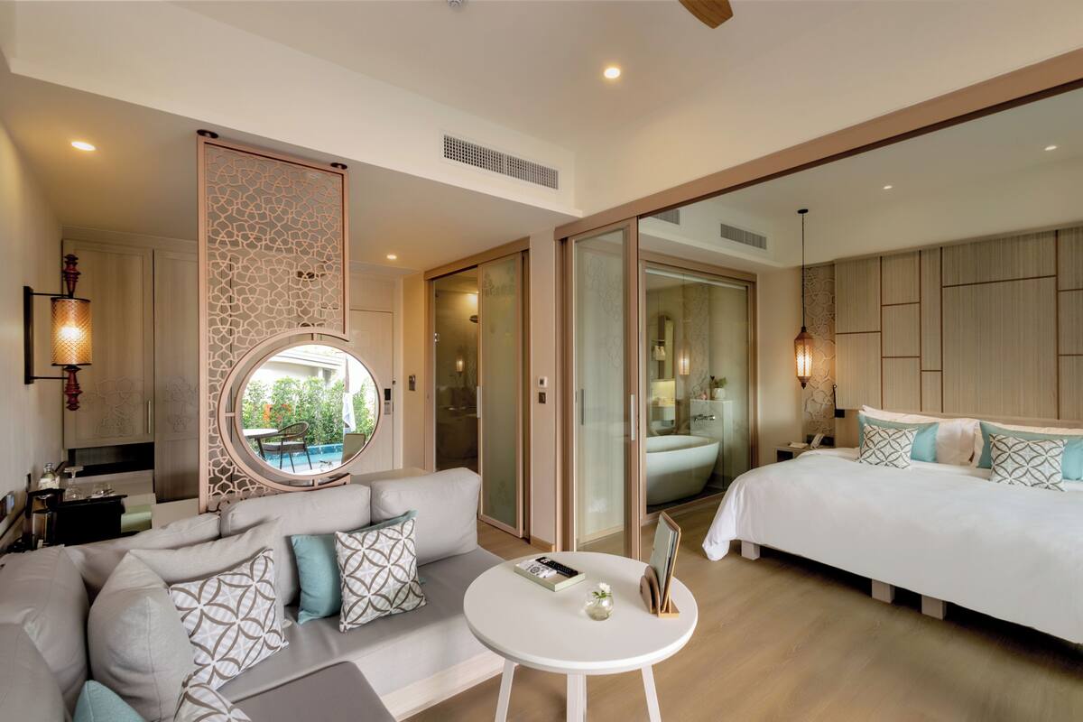 A modern hotel room with a sofa, a small round table, a bed, and a large window.