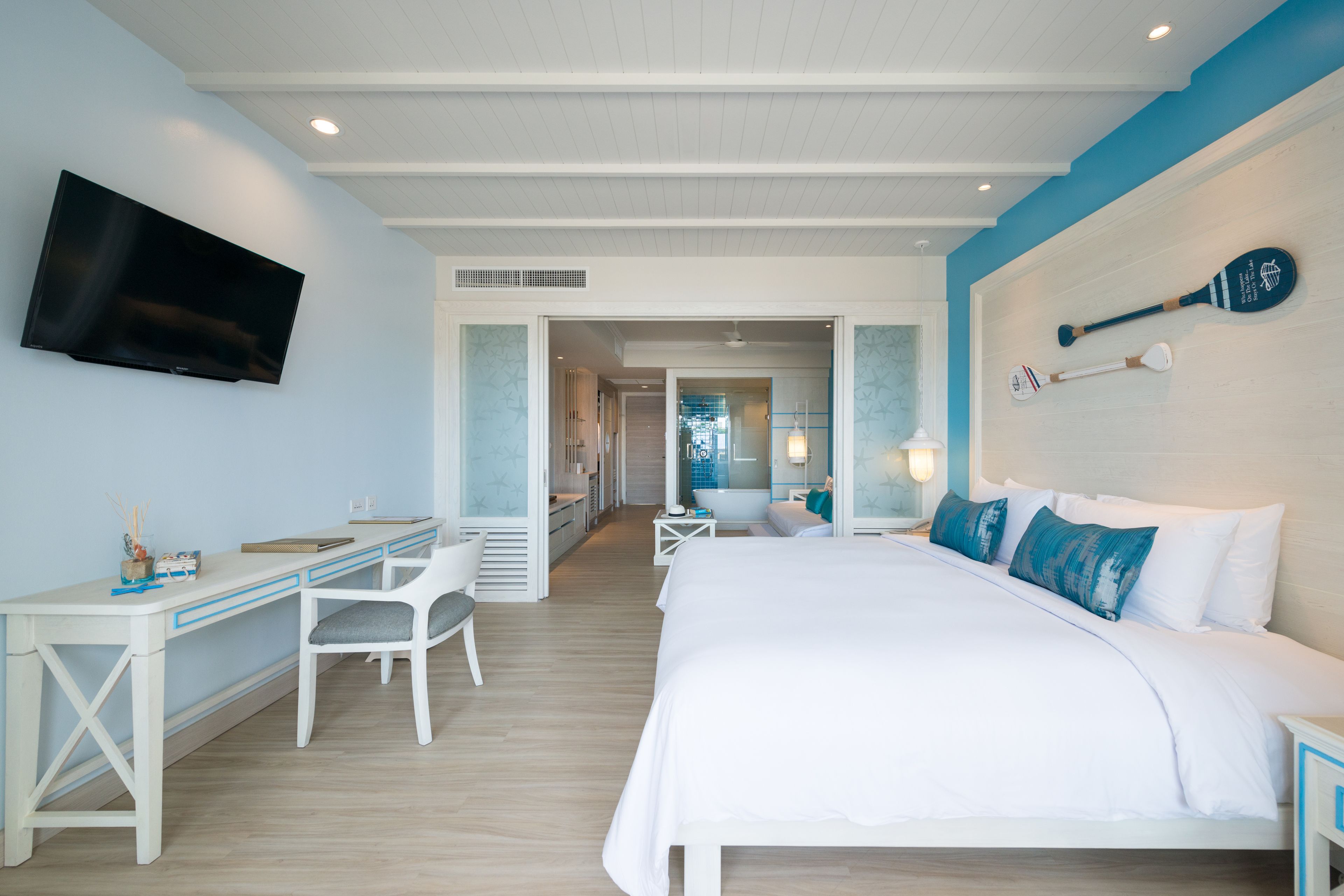 A hotel room with a bed, a desk, a chair, a television, and a wall-mounted paddleboard.