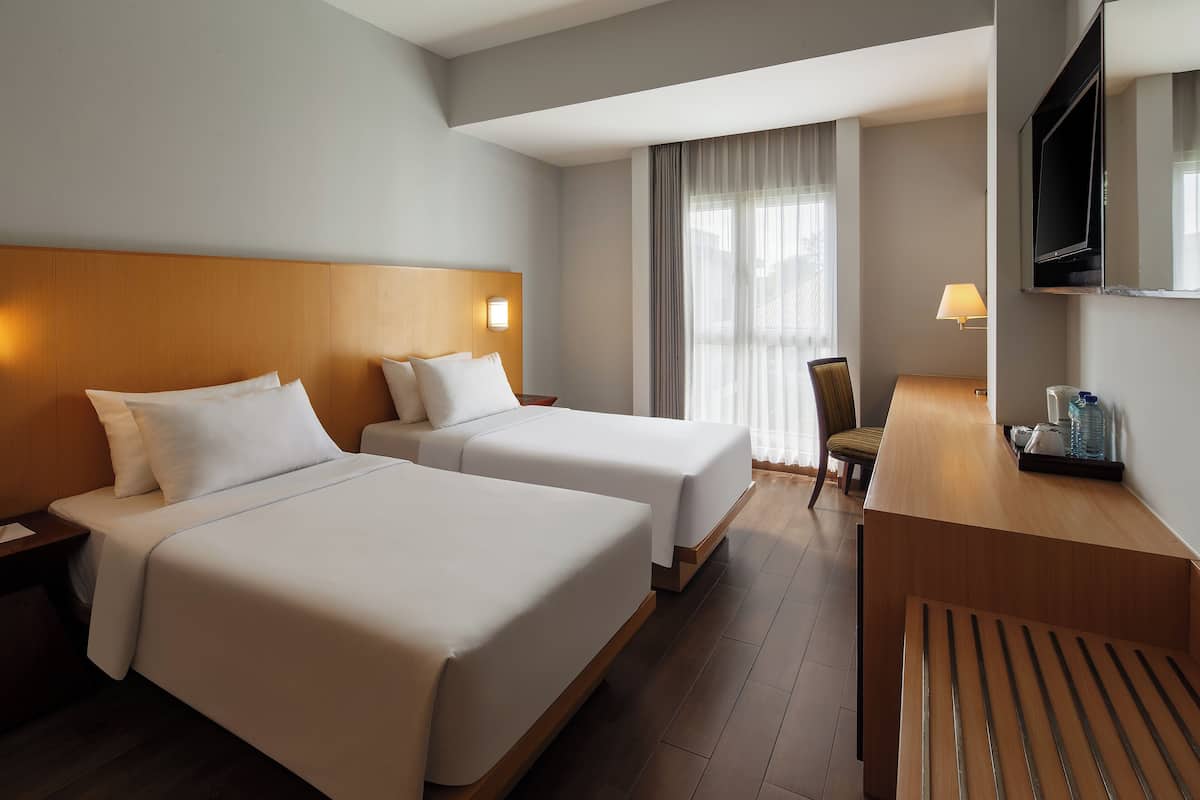 A hotel room with two beds, a desk, a chair, a TV, and a lamp.