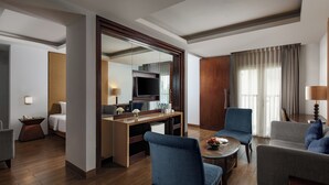 A hotel room with a wooden floor, a desk with a flat-screen TV, a sofa, a chair, and a dining table.