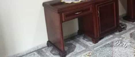 A room with a wooden desk, a mirror, a television mounted on the wall, and a patterned carpet.