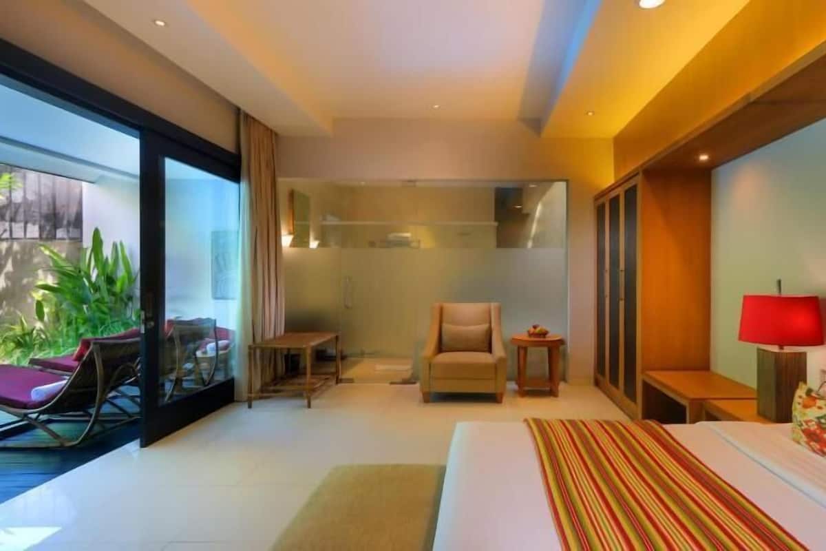 A modern hotel room with a large bed, a sitting area with a chair and table, a small side table, and a view of greenery outside.