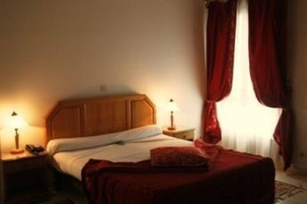 A hotel room with a wooden headboard, a bed with white linens, a red throw blanket, and two bedside lamps.