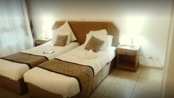 A hotel room with two single beds, each with a pillow and a blanket. There is a nightstand with a lamp on one side and a small table with a phone on the other.