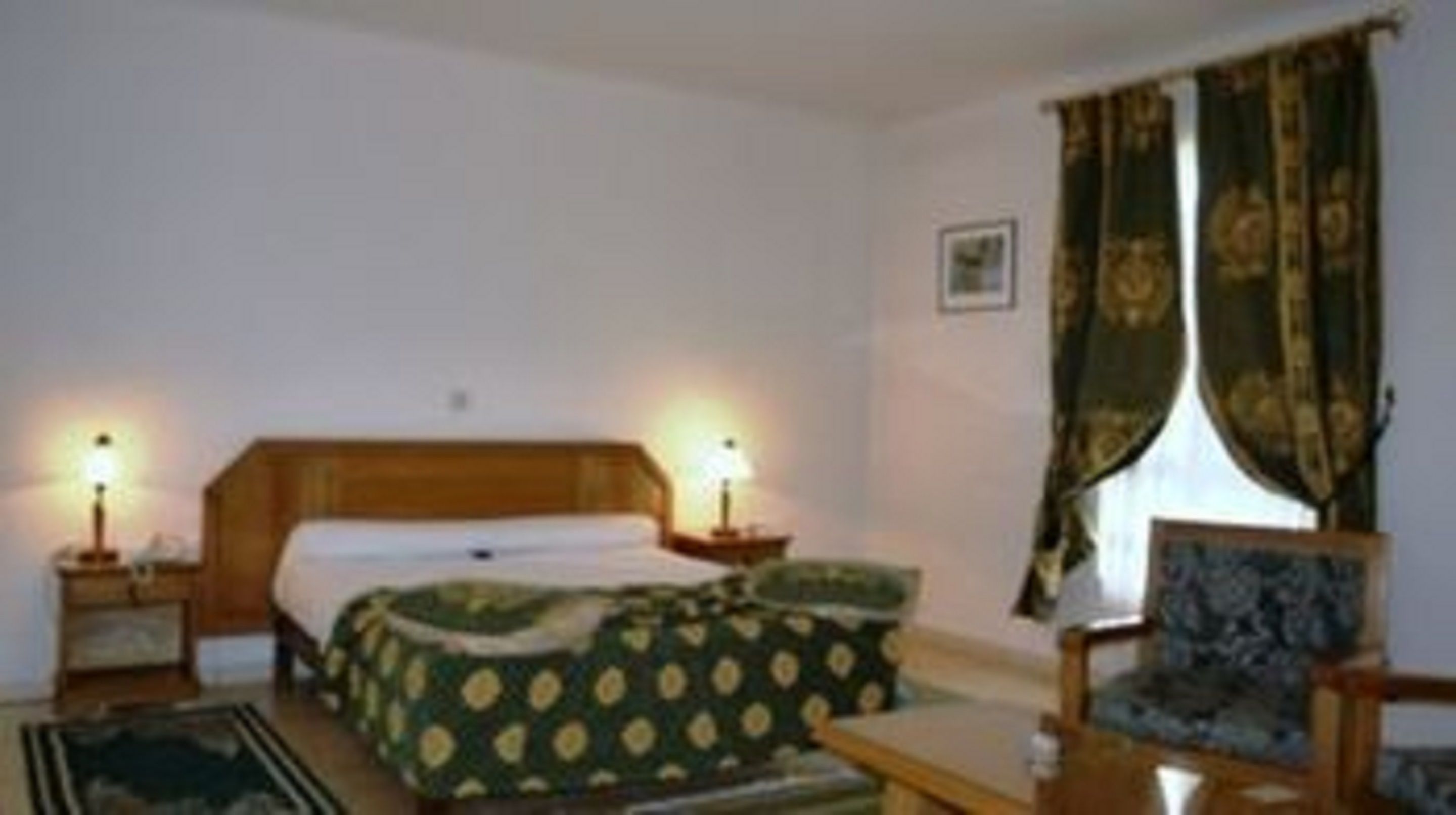 A bedroom with a bed, two bedside tables, a chair, a lamp, and a window with curtains.
