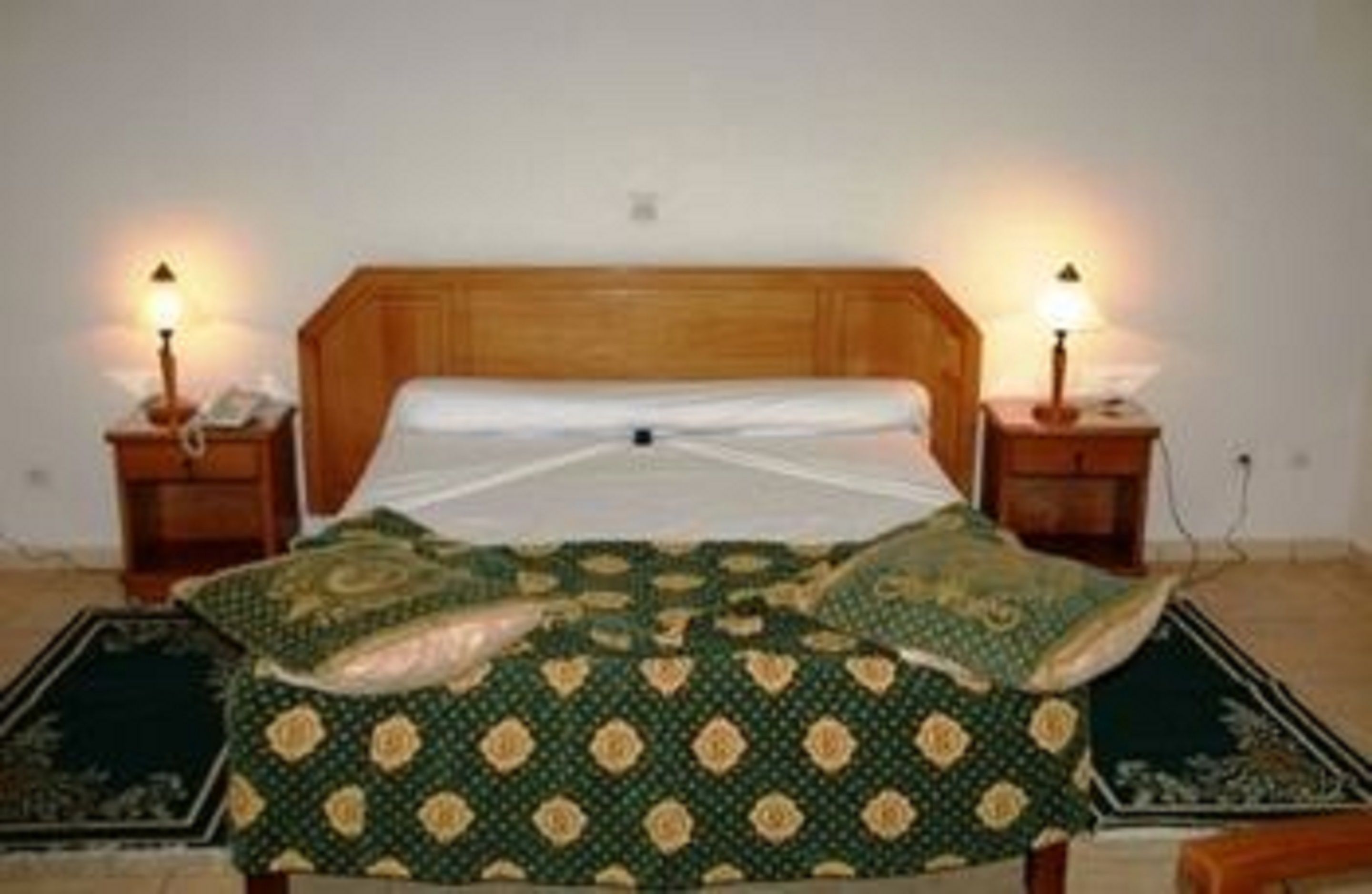 Un dormitorio con cabecera de madera, colcha estampada verde, dos lámparas de noche y una mesita de noche con un teléfono.
