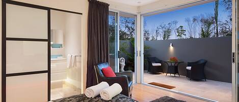 A modern hotel room with a bed, a massage chair, a small table with chairs, and a view of the outdoors.