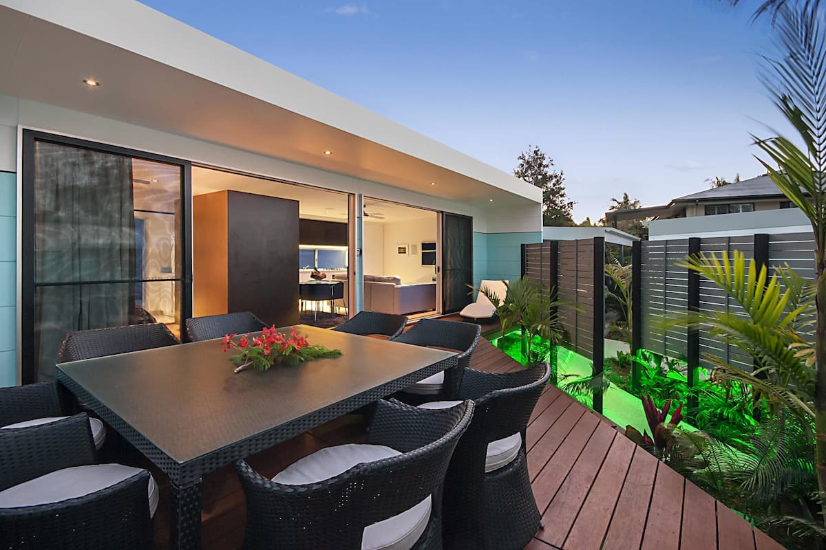 Un espacio al aire libre moderno con comedor, una mesa grande y sillas, rodeado de follaje y una terraza de madera.