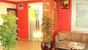 A lobby with a red wall, a beige sofa, a potted plant, and a small round table with books. - Hotel Sweet (Algiers)