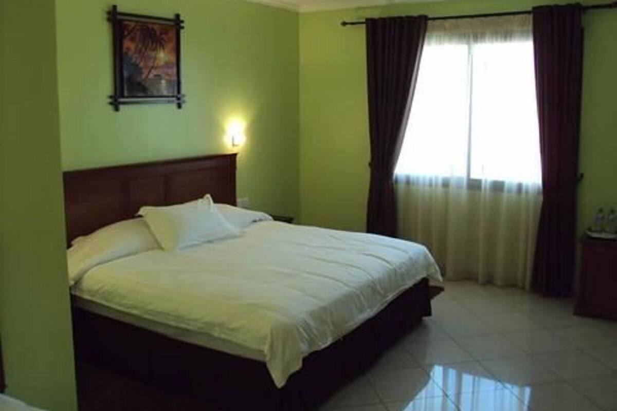 A hotel room with a single bed, a nightstand, a painting on the wall, and a window with curtains.