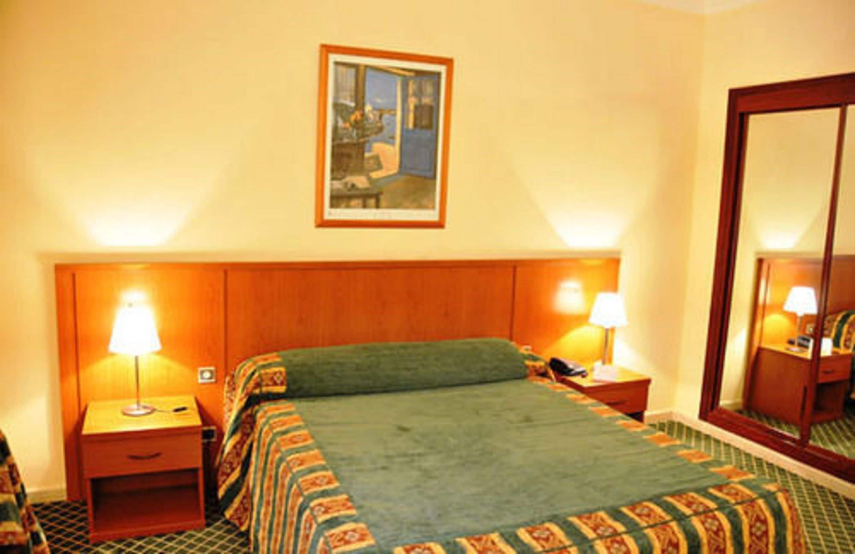 Habitación de hotel con cabecera de madera, dos mesitas de noche con lámparas, cama con manta verde, un cuadro enmarcado en la pared y un espejo grande.