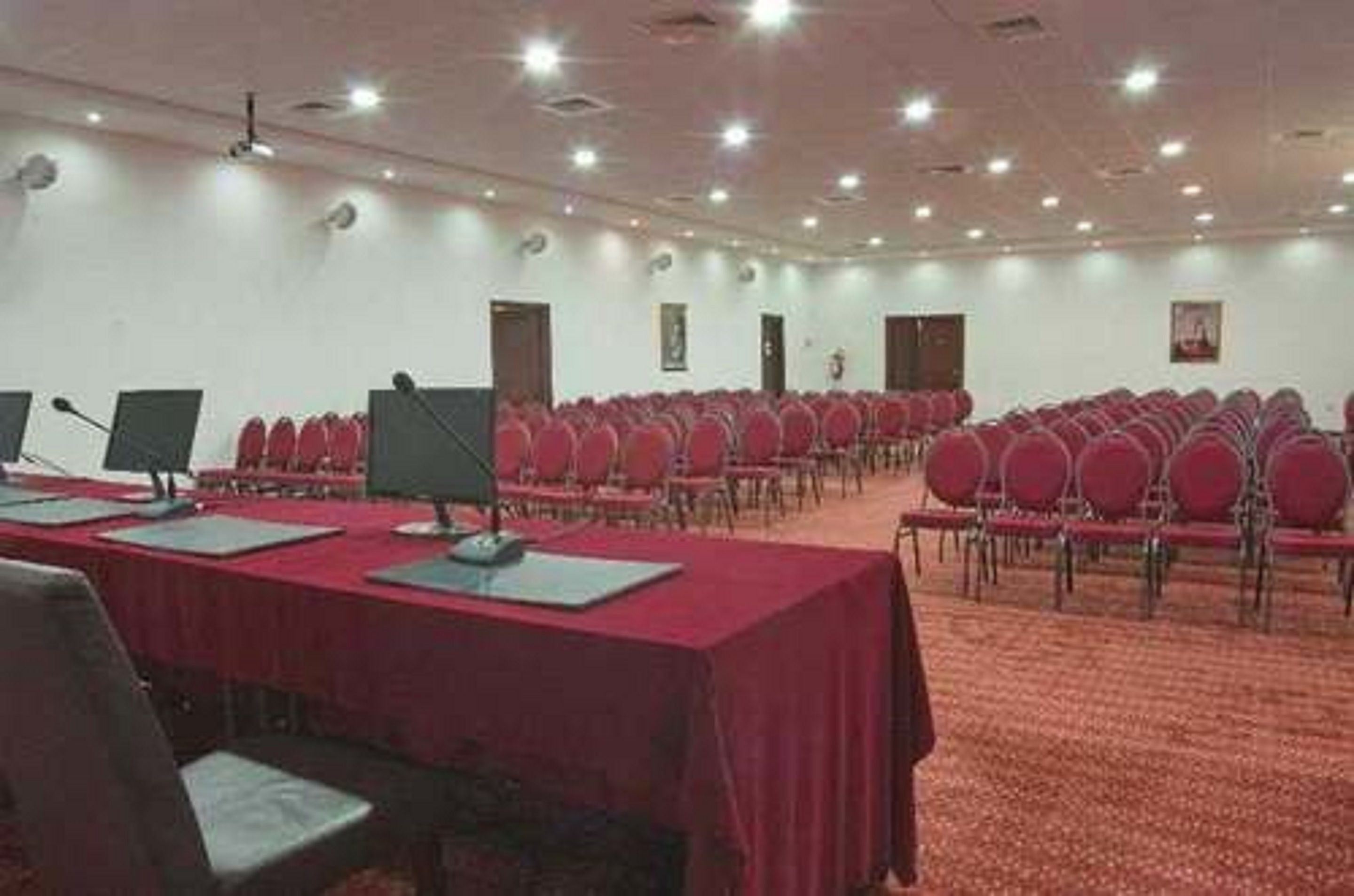 Un salón de conferencias con filas de sillas rojas, una larga mesa con micrófonos y suelo alfombrado.