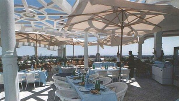 Un espace de restauration en plein air avec des tables et des chaises blanches, protégé par de grands parasols, et offrant une vue sur le paysage urbain en arrière-plan.