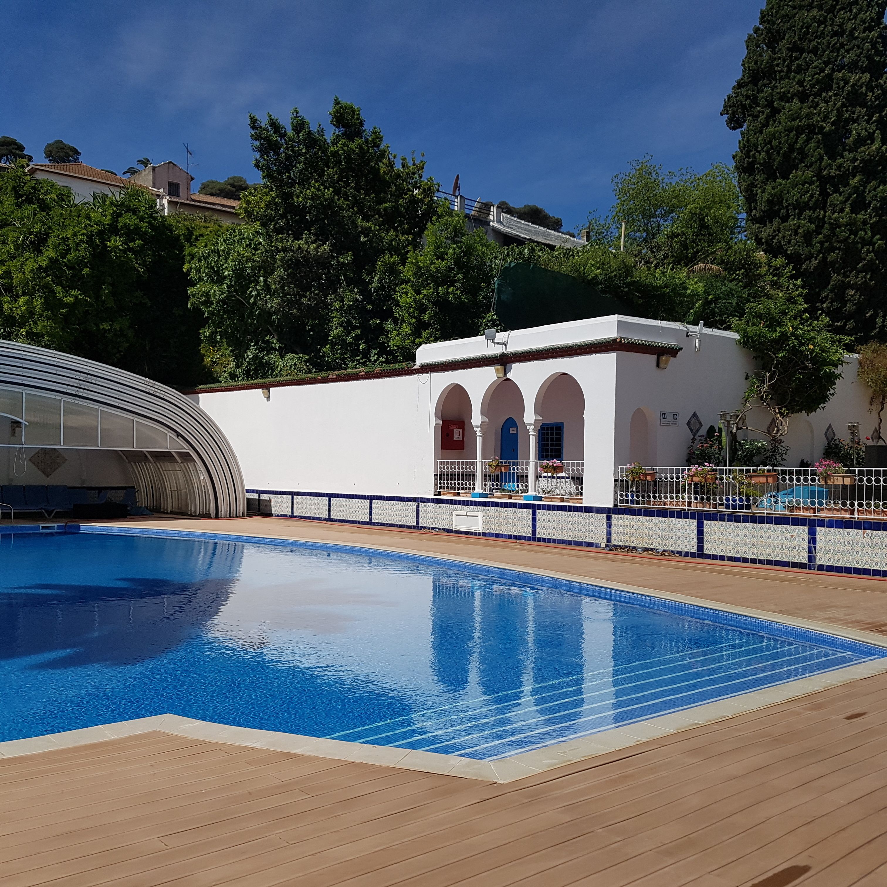 A swimming pool with a wooden deck, a white building with arches, and surrounding greenery.