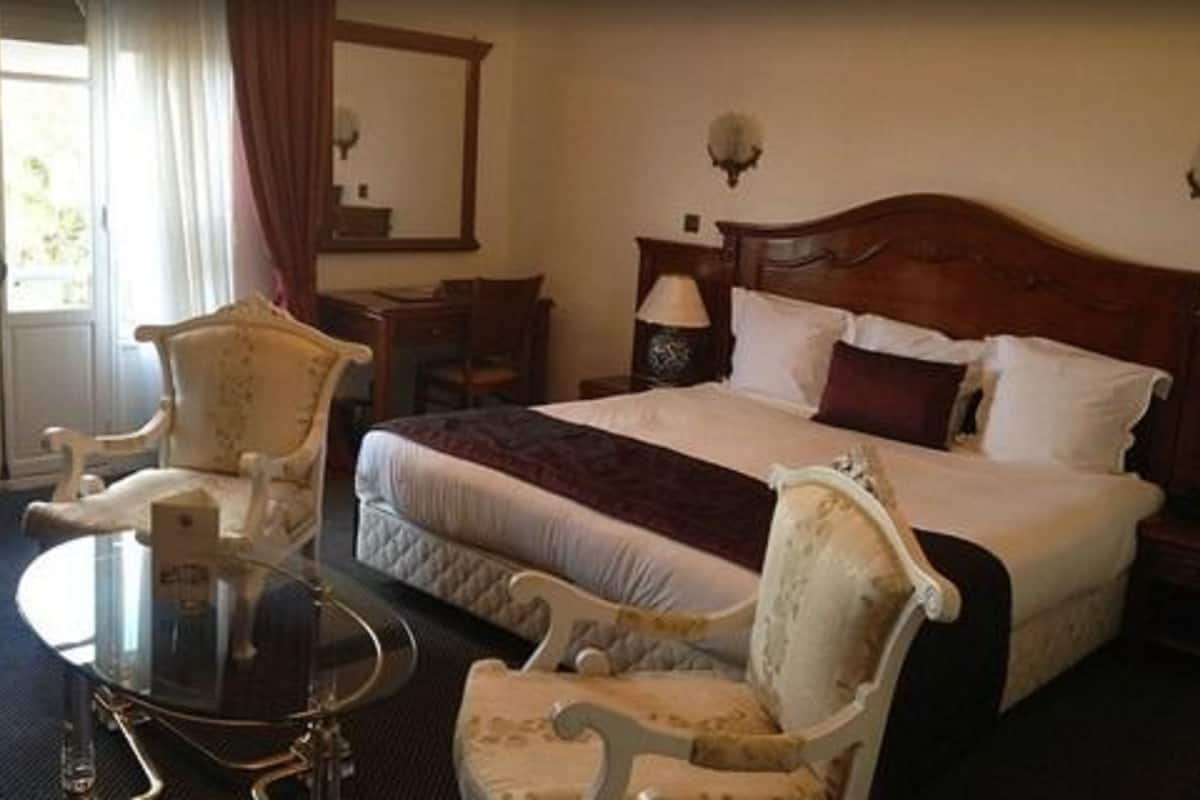 A hotel room with a large bed, two armchairs, a small round table, and a nightstand with a lamp.
