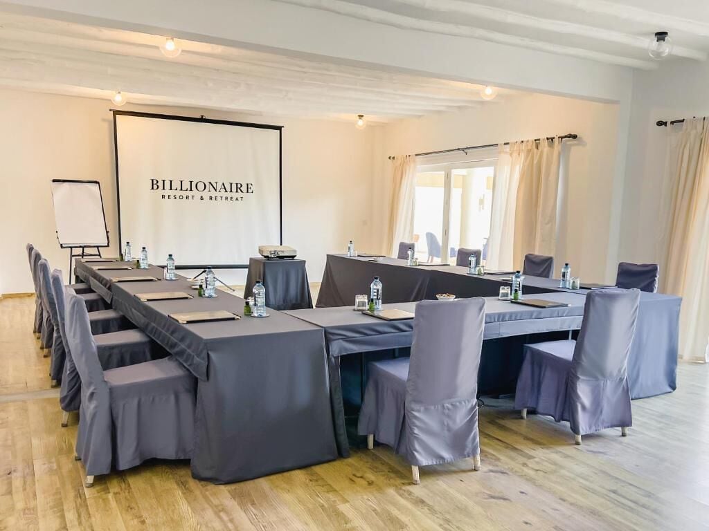 A conference room with a long table, chairs, a projector screen, and a banner reading 'Billionaire Resort & Retreat'.
