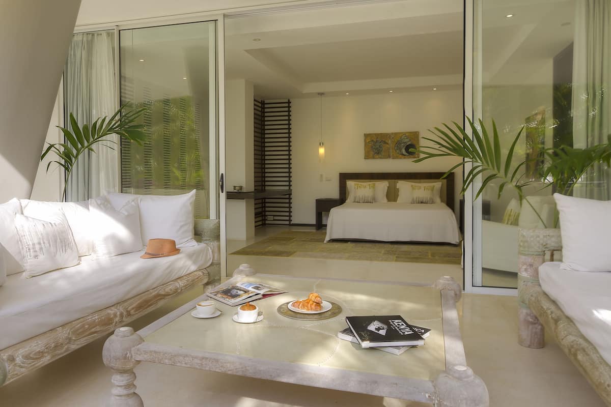 A modern hotel room with a glass door leading to a balcony, a bed with white bedding, a coffee table with a croissant and a magazine, and a sofa with white cushions.
