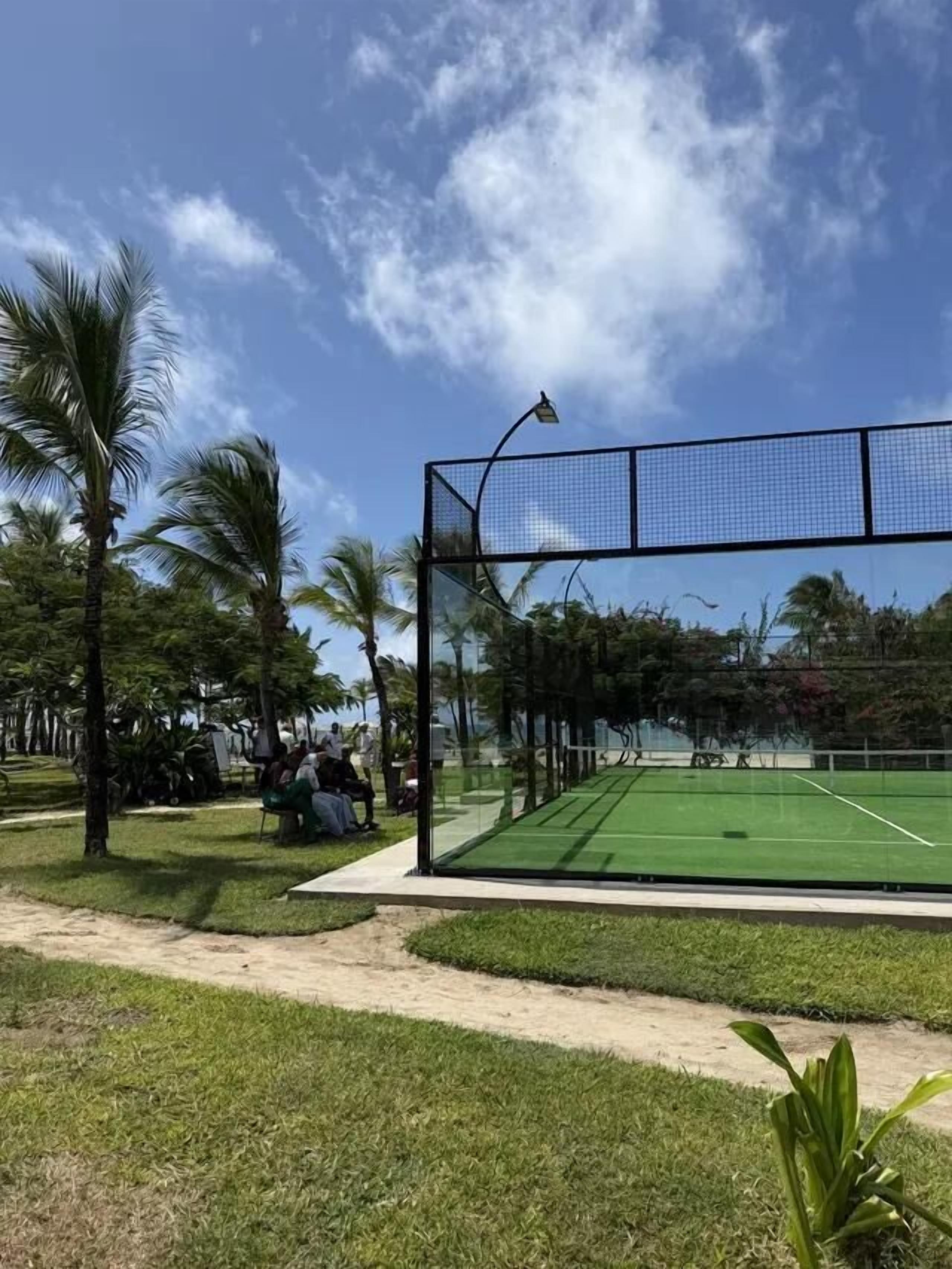 Cancha de tenis al aire libre con red, rodeada por un sendero para caminar y palmeras.