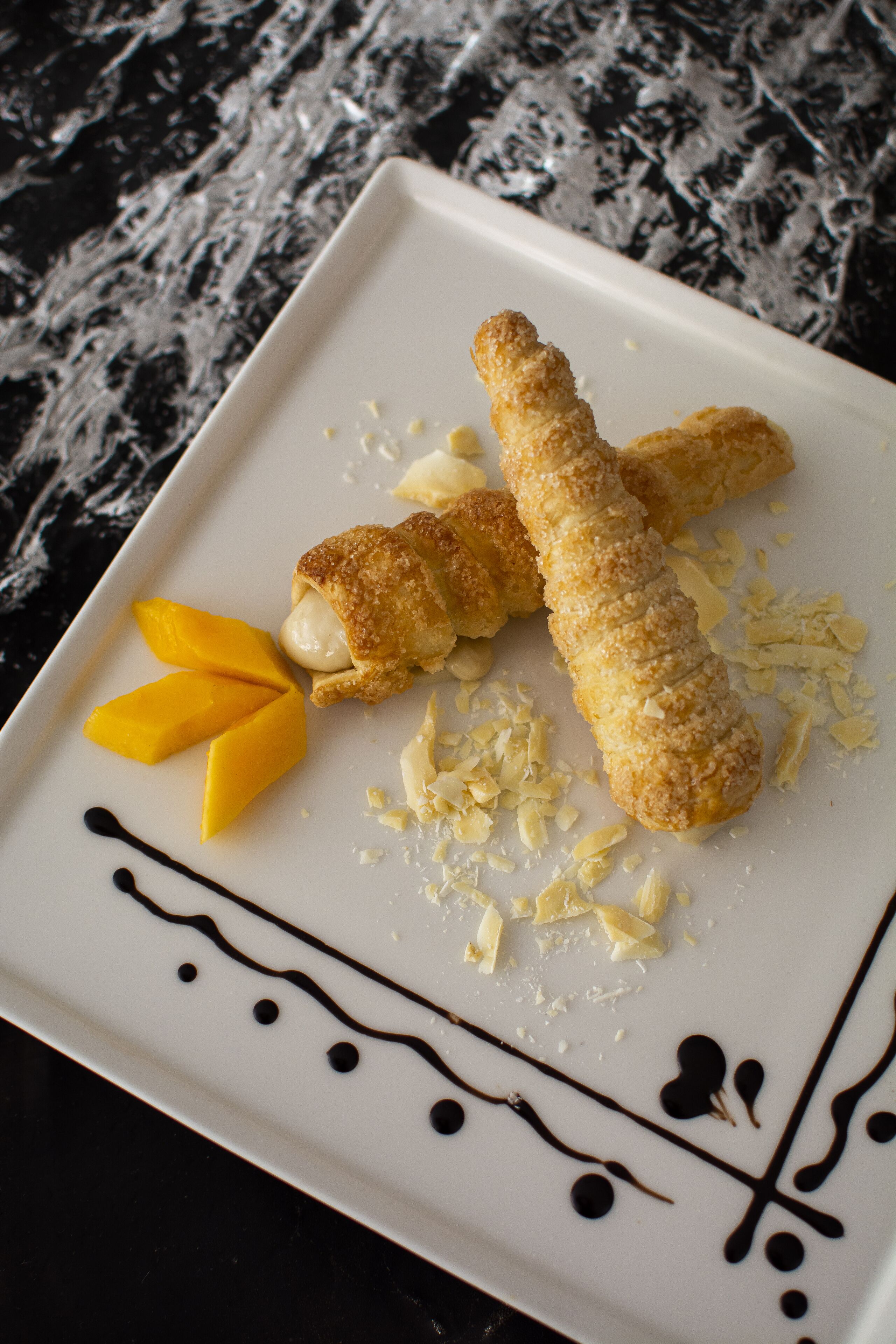 A white plate with two breaded items, mango slices, and chocolate drizzle.