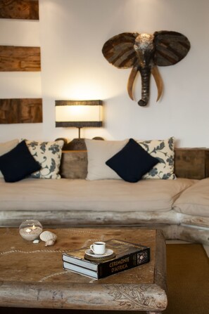 Un salon chaleureux avec un canapé, des coussins décoratifs, une table basse sur laquelle se trouvent un livre et une bougie, et une tête d’éléphant fixée au mur.