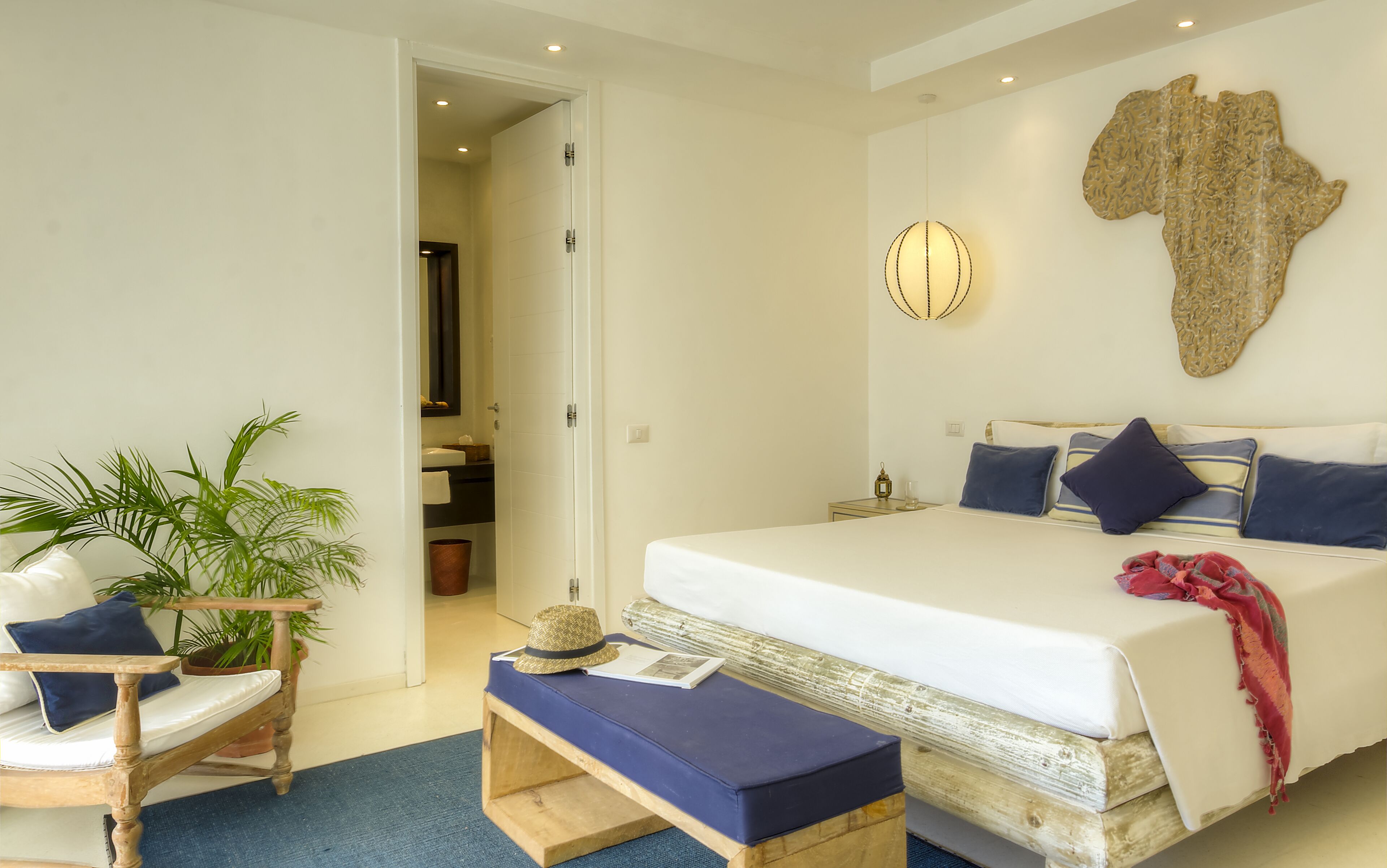 Un dormitorio con una cama, un banco de madera, una silla, una planta en maceta y un aplique de pared.