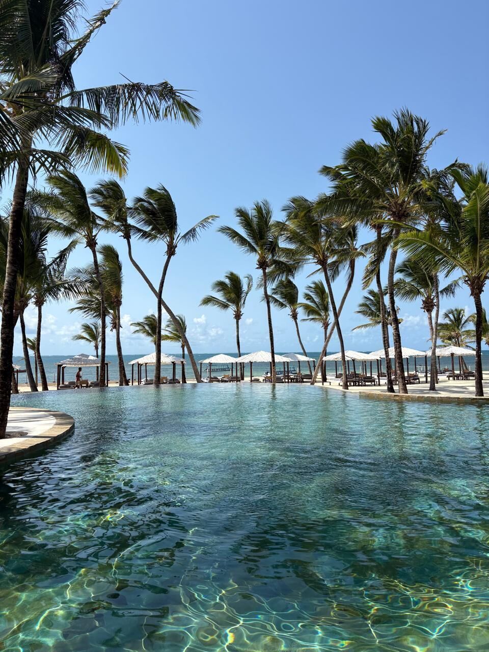 Piscina límpida rodeada por palmeiras e guarda-sóis de praia, com vista para o oceano.