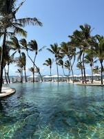 A clear pool surrounded by palm trees and beach umbrellas, with a view of the ocean.