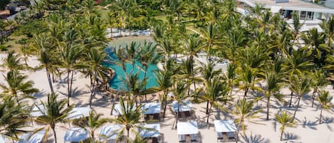An aerial view of a resort with a central swimming pool surrounded by palm trees and beach chairs.