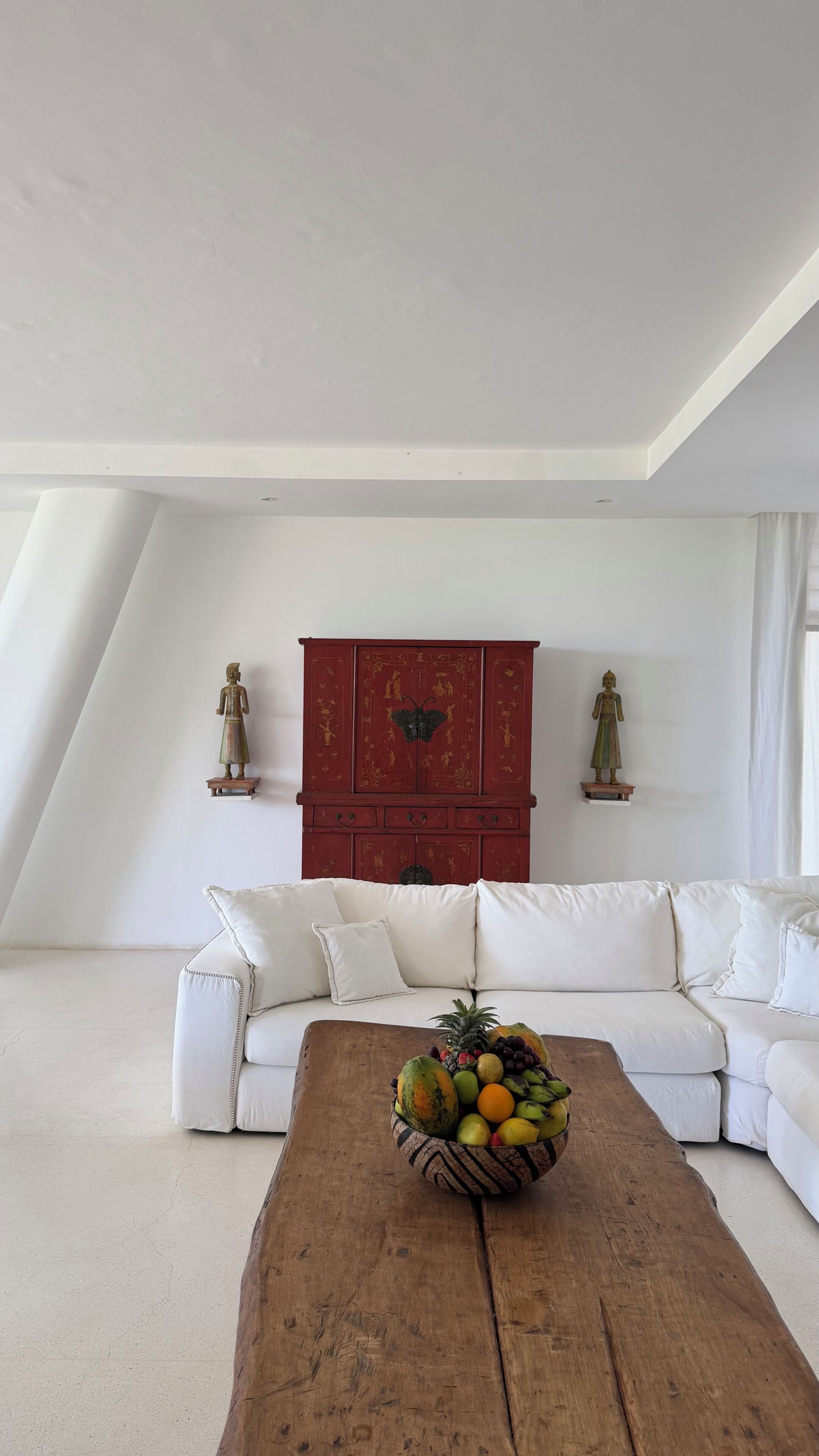 A modern living room with a white sofa, a wooden coffee table with fruit, and a red cabinet with decorative figures.