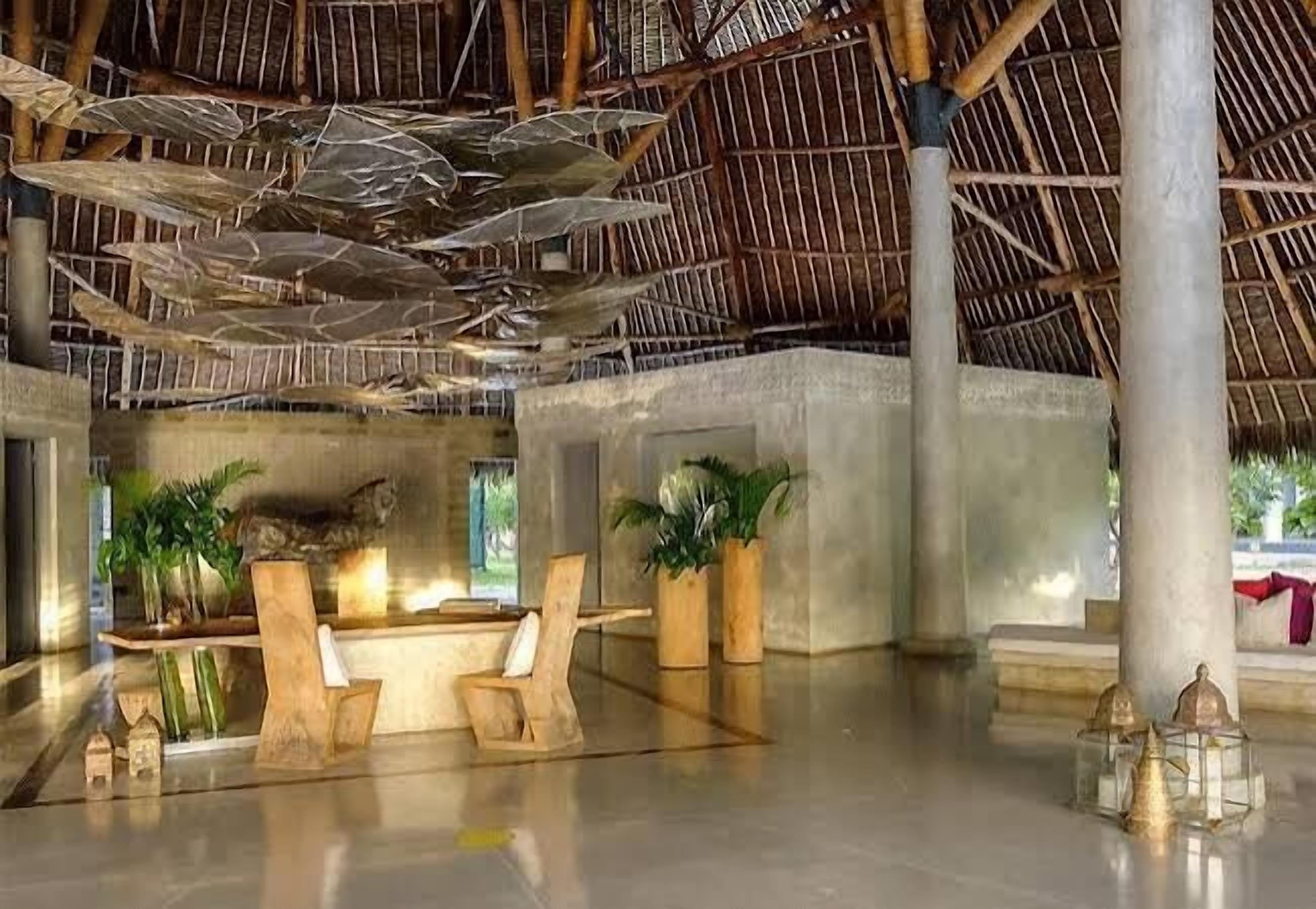 A spacious lobby with a high ceiling, concrete pillars, and a thatched roof. There are wooden chairs and potted plants.