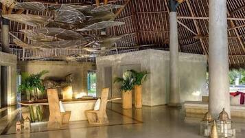 A spacious lobby with a high ceiling, concrete pillars, and a thatched roof. There are wooden chairs and potted plants.
