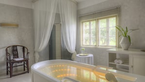 Un baño con bañera, una silla, una mesita con una planta y una ventana con persianas.