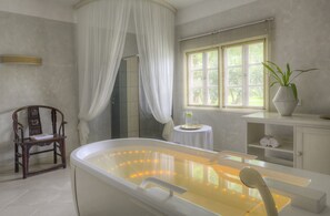 Une salle de bain avec une baignoire, une chaise, une petite table avec une plante et une fenêtre avec des stores.