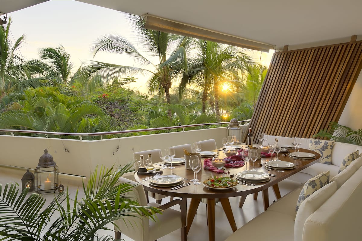 A dining area with a table set for four, surrounded by a balcony with a view of palm trees and greenery.